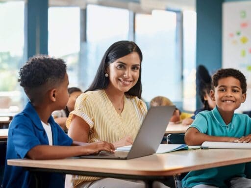 A smiling teacher and two young students working together on laptops and notebooks in a classroom, symbolising the use of notebooklm for students through google workspace for education.